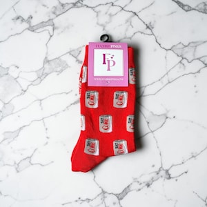 May include: Bright red socks patterned with white and silver Diet Coke cans. The socks have a pink tag with the text "FUCHSIA PINKS" and a logo. The socks are suspended from a black hook.
