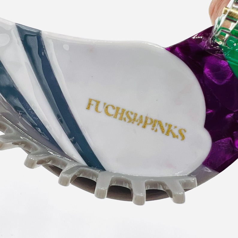 May include: A white and purple hair clip with a gold inscription that reads "FUCHSIA PINKS" and a gray, toothed edge.