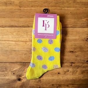 Pode incluir: Um par de meias amarelas com um padrão de bolinhas em azul, roxo e bege. As meias têm uma etiqueta com o texto "FUCHSIA PINKS" e o endereço do site. As meias estão penduradas em um gancho preto.