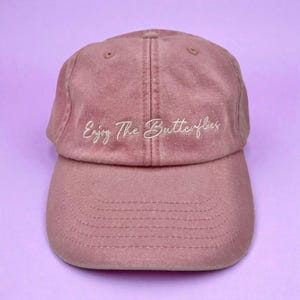 Puede incluir: Gorra de béisbol rosa empolvado con visera curva. La frase "Enjoy The Butterflies" está bordada en blanco. El gorro está hecho de una tela suave y tiene un diseño clásico de seis paneles.