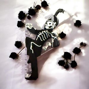 Peut inclure: Une figurine de squelette noir et blanc en uniforme de fanfare. La figurine tient un objet rectangulaire transparent. Des fleurs noires séchées sont éparpillées autour de la figurine sur une surface blanche.