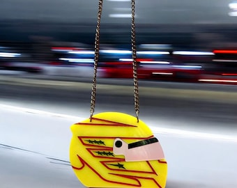Puede incluir: Un collar con forma de casco de carreras amarillo con detalles rojos y verdes. El casco está colgado de una cadena dorada.