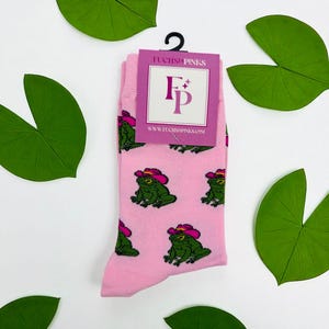 May include: Pink socks with a repeating pattern of green frogs wearing pink cowboy hats. The socks have a tag that reads "FUCHSIA PINKS" and "www.fuchsiapinks.com". The socks are surrounded by green lily pads.
