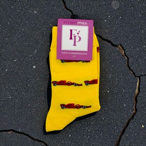 May include: Yellow socks with a red race car pattern. The socks have black heels and toes. A purple tag at the top reads "FUCHS&PINKS" and "www.FUCHSPINKS.com". The tag also features a stylized "F" and "P" logo.