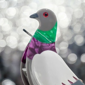 May include: A white, gray, green, and purple ceramic dove figurine with a red eye. The dove is perched on a small, gray base.