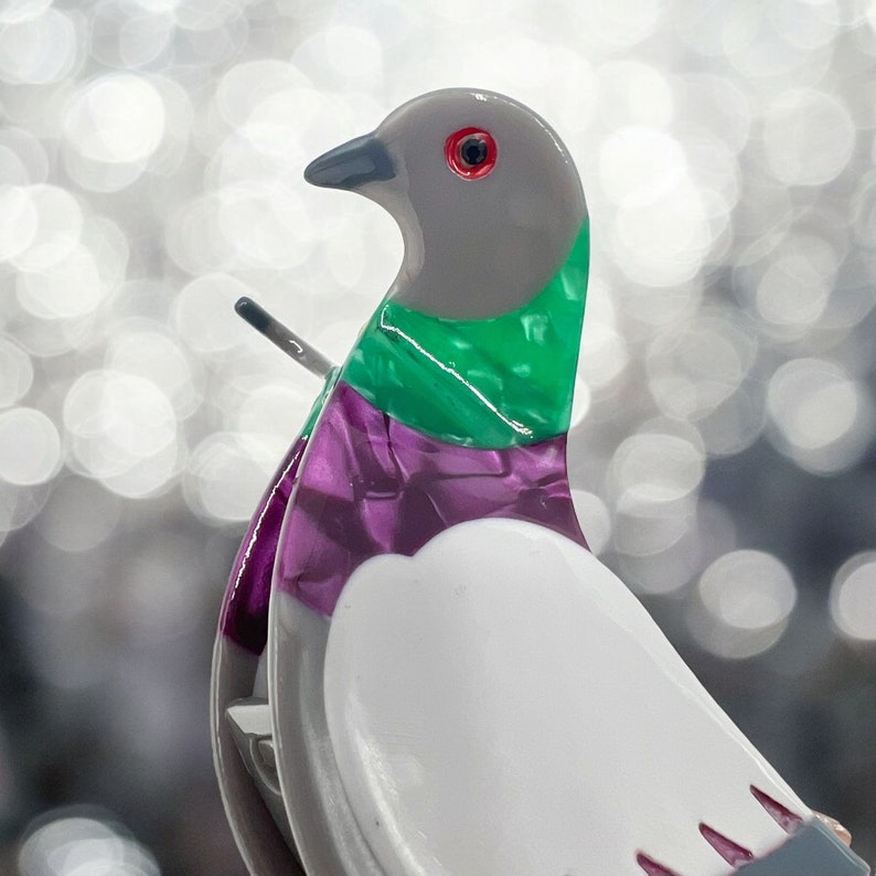 May include: A white, gray, green, and purple ceramic dove figurine with a red eye. The dove is perched on a small, gray base.
