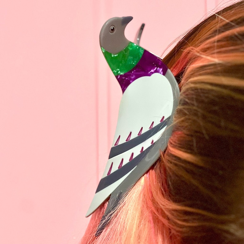 May include: A white, gray, and purple bird hair clip with green accents. The bird is perched on a person's hair.