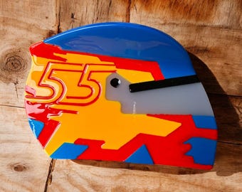 Puede incluir: Un colorido reloj de pared con forma de casco de carreras. El reloj presenta una parte superior azul, detalles rojos y amarillos, y el número "55" en una fuente audaz. El reloj tiene una visera gris y una manecilla negra.