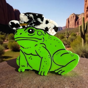 May include: A vibrant green frog brooch, styled with a black and white cowboy hat. The frog has black outlines and is set against a desert backdrop with cacti and red rock formations. A playful, unique accessory.