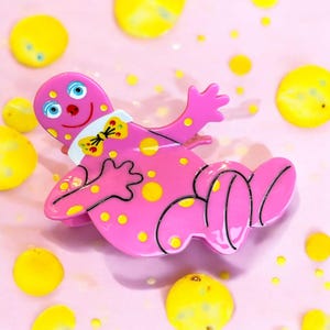 May include: Pink plastic hair clip shaped like a cartoon character with yellow polka dots and a bow tie. The character has a smiling face with blue eyes.