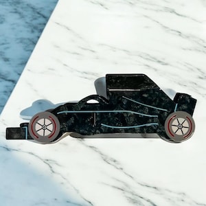 May include: A black race car-shaped brooch with a textured surface and light blue accents. The wheels are gray with red and black details. The brooch is set against a white and gray marble background.