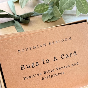 May include: A stack of rectangular kraft paper cards inside a matching cardboard box tied with a green ribbon. The top card features black printed text that reads: BOHEMIAN REBLOOM, Hugs In A Card, Positive Bible Verses and Scriptures.