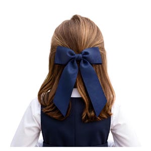 May include: A large, solid navy blue hair bow made of grosgrain ribbon with two long, pointed tails. The bow is secured at the center with a matching fabric knot and is positioned at the back of the head, resting against light brown hair.