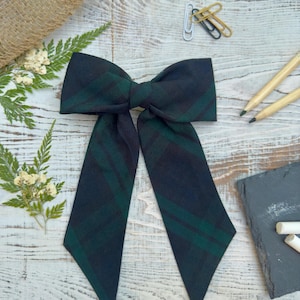 May include: A green and black plaid bow with long tails. The bow is tied in a knot and the tails are hanging down. The bow is on a wooden surface.