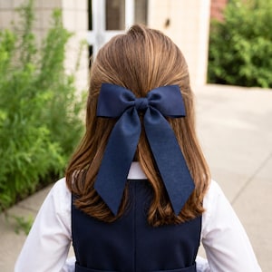 May include: A navy blue hair bow with long streamers is tied in the hair of a person wearing a white shirt and navy blue jumper. The bow is the focal point, with the person's hair and clothing providing a contrasting background.