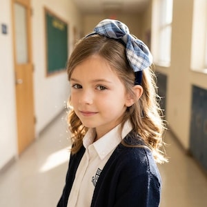 May include: A blue and white plaid fabric bow headband featuring a large, structured bow design. The headband is made of a soft, woven material with a classic checkered pattern in shades of navy, light blue, and white, secured on a dark blue band.