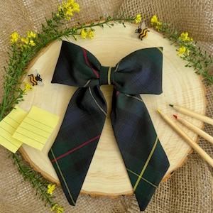 Puede incluir: Una pajarita de tartán verde oscuro con rayas rojas y amarillas. La pajarita está sobre una rodaja de madera con flores amarillas, lápices y decoraciones de abejas.