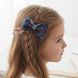 May include: A hair bow featuring a dark blue and grey plaid pattern. The bow is constructed with a structured fabric and is attached to a clip, secured in long, light brown hair that is styled with a side braid detail on the side of the head.