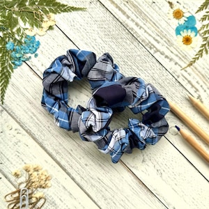 May include: Two blue and white plaid scrunchies with a navy blue center. The scrunchies are on a white wooden surface.