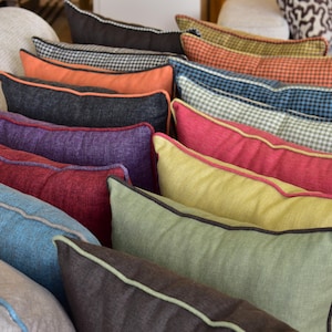 May include: A collection of rectangular throw pillows in various solid colors and patterns, including blue, purple, red, yellow, green, orange, and black. Each pillow features a contrasting piped edge along the seams, creating a structured look.