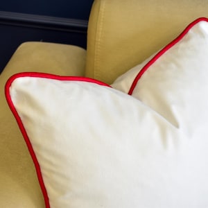 May include: Close-up of two white throw pillows with red piping. The pillows are on a light yellow sofa. The background includes a dark blue wall and a portion of the sofa.