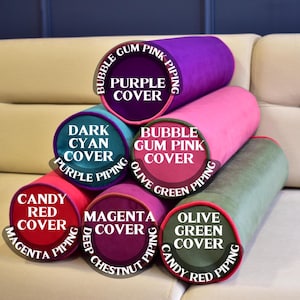 May include: A stack of cylindrical pillows in various colors, including purple, pink, green, and red. Each pillow has a contrasting color piping and a label indicating the cover and piping colors. The pillows are arranged on a beige sofa.