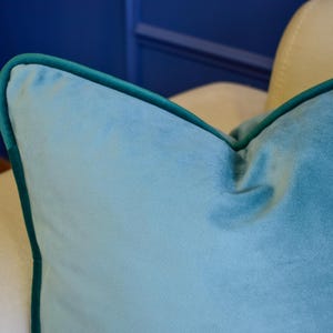 May include: Close-up of a teal velvet pillow with a dark green trim. The pillow is on a cream-colored chair, with a blue wall in the background. The pillow's texture is soft and plush.