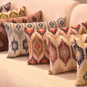 May include: A collection of decorative throw pillows with patterned designs in various colors, including red, brown, blue, and yellow. The pillows are arranged on a cream-colored sofa, showcasing their intricate patterns and textures.