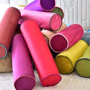 Könnte beinhalten: Eine Sammlung bunter zylindrischer Kissen mit kontrastierenden Einfassungen. Die Kissen sind aus einem weichen, samtigen Stoff gefertigt und liegen auf einer weißen Oberfläche in einem Stapel.