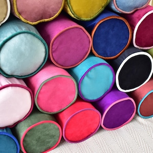 May include: A collection of colorful, cylindrical bolster pillows in various shades of pink, blue, green, purple, and yellow. Each pillow has a contrasting trim, creating a visually appealing display. The pillows are stacked together, showcasing their soft, plush texture.