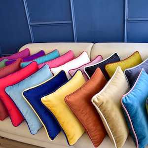 Puede incluir: Una colección de cojines coloridos colocados en un sofá beige. Los cojines son de varios tonos, incluyendo rojo, azul, amarillo y verde, con ribetes contrastantes. Los cojines son rectangulares con una forma única.