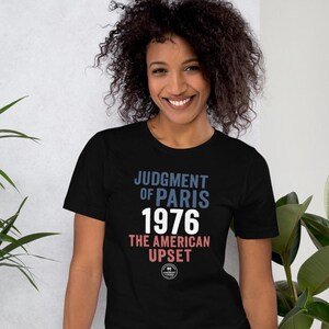 Puede incluir: Camiseta negra con el texto "JUDGMENT OF PARIS 1976 THE AMERICAN UPSET" en letras azules y blancas. La persona que lleva la camiseta tiene el pelo rizado oscuro y sonríe.