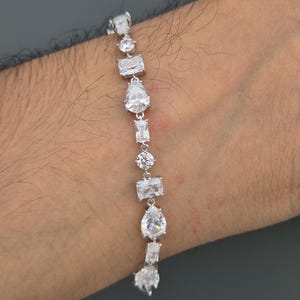 Könnte beinhalten: Ein silbernes Armband mit einer Reihe klarer, funkelnder Edelsteine in verschiedenen Formen, darunter rund, rechteckig und tropfenförmig. Das Armband wird vor einem neutralen Hintergrund präsentiert, um die Brillanz der Steine hervorzuheben.