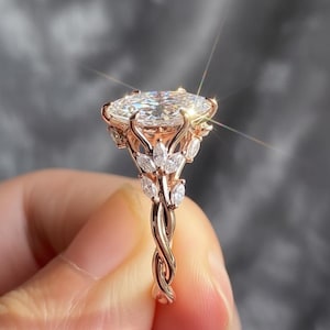 May include: Rose gold ring with a large, oval-cut diamond. The ring has a floral design with smaller diamonds and a twisted band. The ring is held up, showing its sparkle against a blurred background.