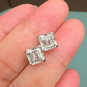 Op de afbeelding: Een paar vierkant geslepen diamanten oorstekers. De diamanten zijn gezet in een zilveren of witgouden zetting. De oorbellen worden in een hand gehouden tegen een lichtblauwe achtergrond.