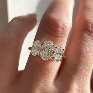 May include: A gold ring featuring three oval-cut diamonds. The diamonds are set closely together, creating a continuous sparkle. The ring is shown on a finger, catching the light and showcasing the brilliance of the stones.