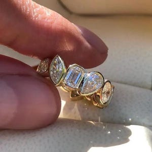 May include: A gold ring featuring a series of differently shaped clear gemstones. The gemstones include marquise, emerald, and pear cuts. The ring is held between two fingers.