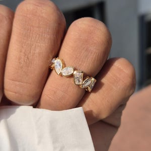 May include: A gold ring featuring a sequence of clear, differently shaped stones. The ring has marquise, oval, and rectangular cut stones set in a gold band. The ring is shown on a finger.