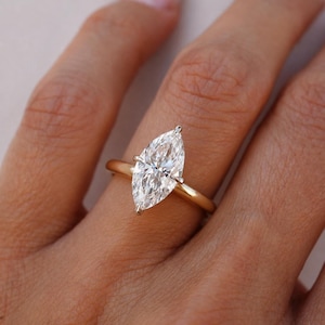 May include: A marquise-cut clear gemstone ring set on a polished yellow gold band. The elongated, pointed stone is secured by a simple metal prong setting, showcasing a brilliant faceted surface that reflects light across its symmetrical shape.