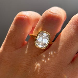 May include: A gold ring featuring a large, rectangular-cut diamond. The diamond is set in a bezel setting, and the ring's band is a simple, polished gold. The diamond sparkles, reflecting light. The ring is a piece of jewelry.