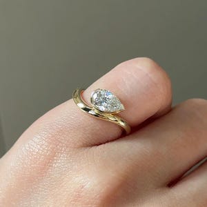 Könnte beinhalten: Ein goldener Ring mit einem birnenförmigen Diamanten. Der Diamant ist in einer Zargenfassung gefasst, und das Band umschlingt den Finger. Der Ring ist ein zartes und elegantes Schmuckstück.
