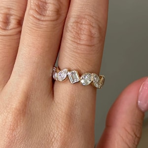Könnte beinhalten: Ein goldener Bandring mit einer Reihe von Diamanten in verschiedenen Formen, darunter rund, birnenförmig und Smaragd. Der Ring ist mit klaren, funkelnden Diamanten besetzt. Der Ring wird an einem Finger getragen.