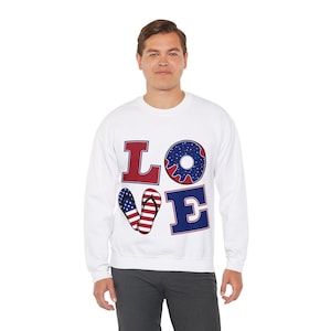 May include: White sweatshirt featuring the word "LOVE" in red and blue. The "O" is a blue donut shape with white stars. The "V" is a pair of flip-flops with a red and white striped design and blue stars.
