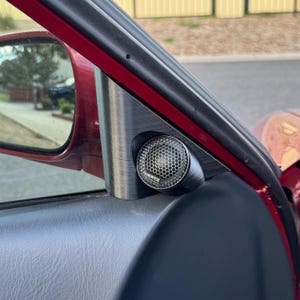 May include: A car interior detail featuring a speaker with a honeycomb grille. The speaker is set in a dark grey, textured panel, contrasted by a red car door frame. The side mirror is visible in the background.
