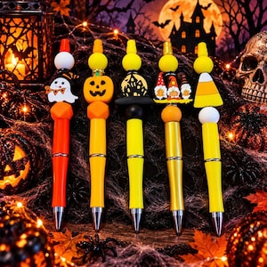 May include: Five Halloween-themed pens featuring colorful silicone toppers. The designs include a white ghost, an orange jack-o'-lantern, a black haunted house, three gnomes holding pumpkins, and a yellow, orange, and white candy corn shape.