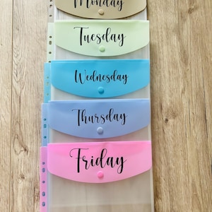 May include: Five clear plastic folders with coloured pockets labelled Monday, Tuesday, Wednesday, Thursday, and Friday. The folders are arranged vertically and have a hole punch on the left side.