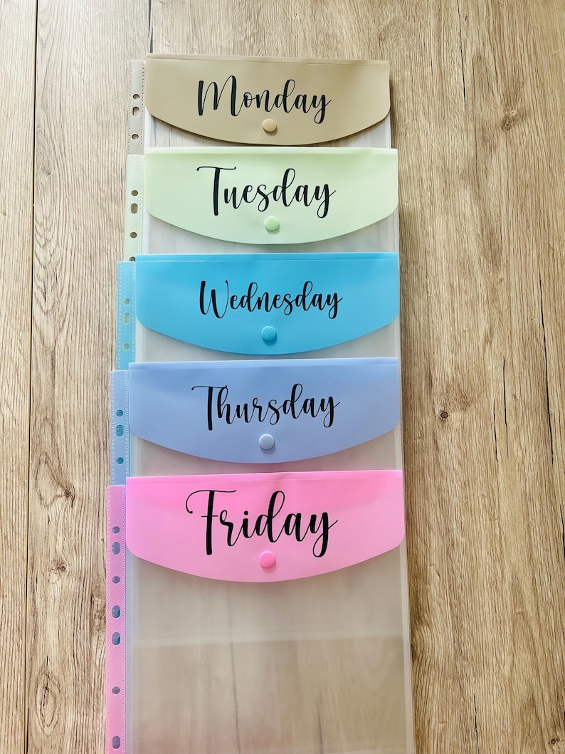 May include: Five clear plastic folders with coloured pockets labelled Monday, Tuesday, Wednesday, Thursday, and Friday. The folders are arranged vertically and have a hole punch on the left side.