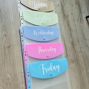 May include: Five clear plastic folders with different coloured pockets and white text. The folders are labelled "Monday", "Tuesday", "Wednesday", "Thursday", and "Friday".