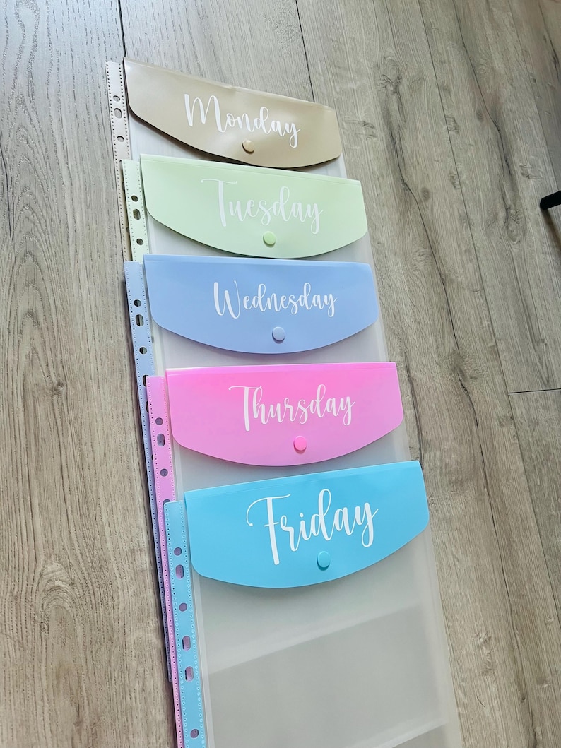 May include: Five clear plastic folders with different coloured pockets and white text. The folders are labelled "Monday", "Tuesday", "Wednesday", "Thursday", and "Friday".