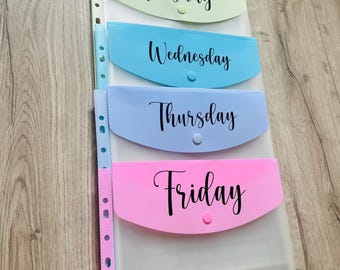May include: A set of five clear plastic folders with colored pockets labeled "Monday", "Tuesday", "Wednesday", "Thursday", and "Friday".